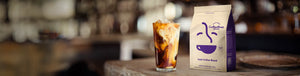 Iced Coffee Blend featured hero -- tall glass of iced coffee with cream and ice cubes resting on table next to Coffee Bean Direct bag
