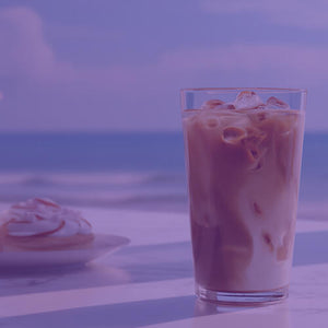Toasted Almond Cream featured mobile hero -- glass of iced coffee and a plate of pastries on a table with a scenic view of the ocean and beach in the background