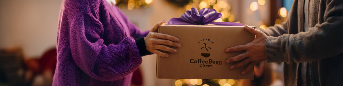 Gift Subscriptions featured hero -- Person giving a Coffee Bean Direct gift box with a purple ribbon to another