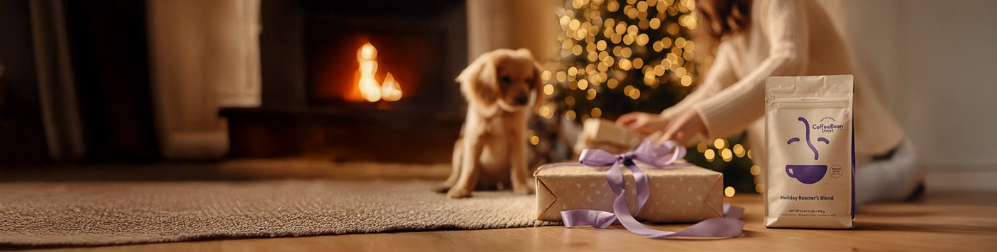 Holiday Roaster's Blend featured hero -- Holiday Roaster's Blend coffee bag and Christmas gift in the foreground with a woman opening a present alongside a puppy and Christmas tree in the background