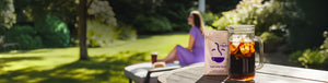 Iced Coffee Blend featured hero -- bag of Coffee Bean Direct Iced Coffee Blend alongside pitcher of iced coffee on a table in the foreground, with a woman in the background enjoying a glass
