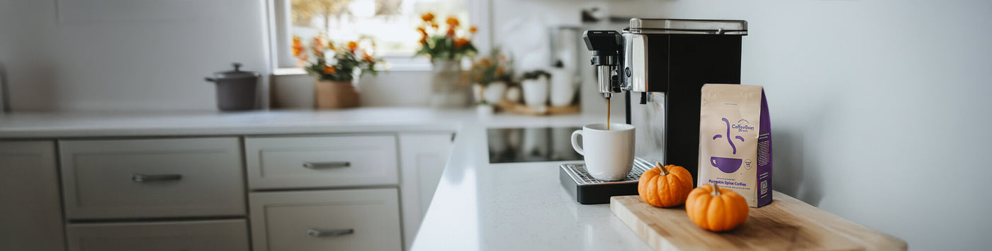 Pumpkin Spice featured hero -- kitchen counter with coffee maker, coffee mug, and small pumpkins on a wooden cutting board alongside a bag of coffee