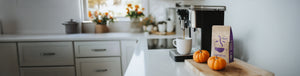 Pumpkin Spice featured hero -- kitchen counter with coffee maker, coffee mug, and small pumpkins on a wooden cutting board alongside a bag of coffee