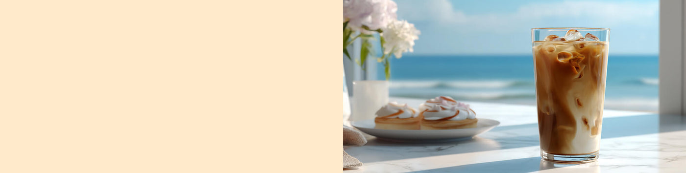 Toasted Almond Cream featured hero -- glass of iced coffee and a plate of pastries on a table with a scenic view of the ocean and beach in the background