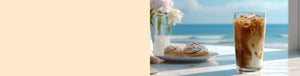 Toasted Almond Cream featured hero -- glass of iced coffee and a plate of pastries on a table with a scenic view of the ocean and beach in the background